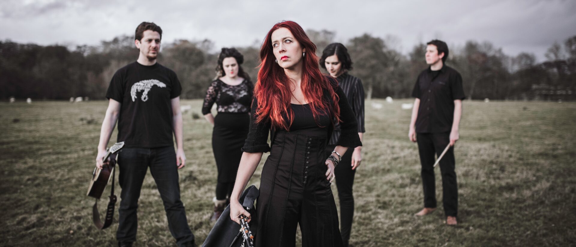 A red-haired woman dressed casually in black and holding the Northumberland pipes poses in a field next to her band mates