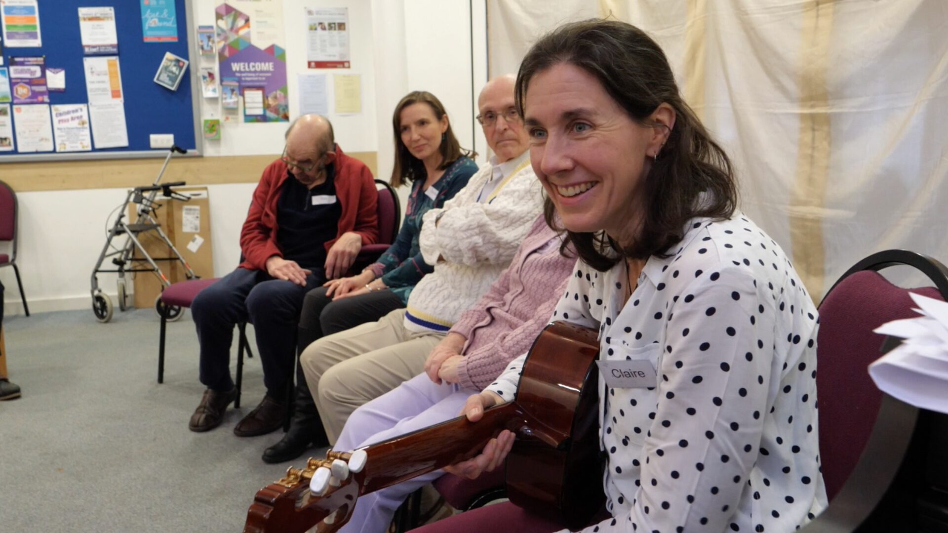 A music therapy group sits making music together, with Claire Molyneux leading them holding a guitar