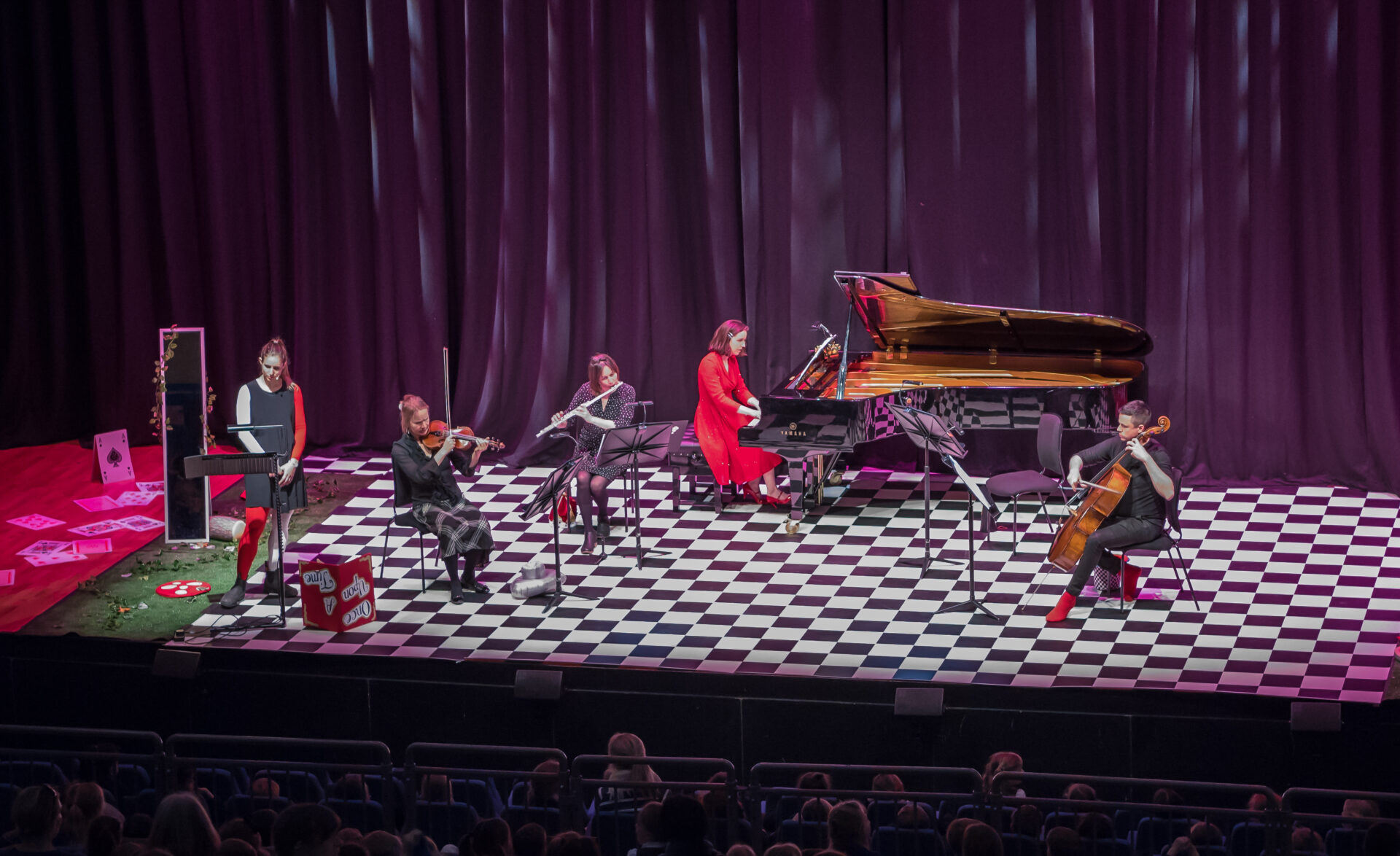 A group of musicians dressed in red and black play on a stage with a chequerboard-patterned floor