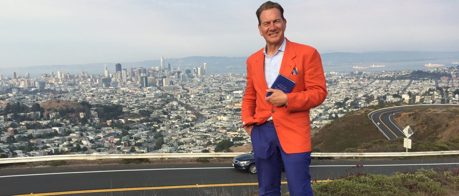 Michael Portillo standing on a hill overlooking a city with a book smiling at the camera.
