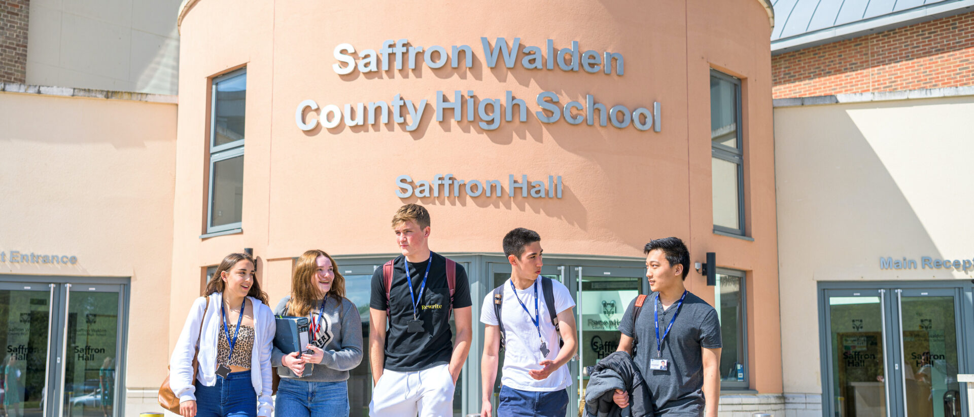 A group of 5 students in casual clothes chat outside the front entrance of Saffron Walden County High School