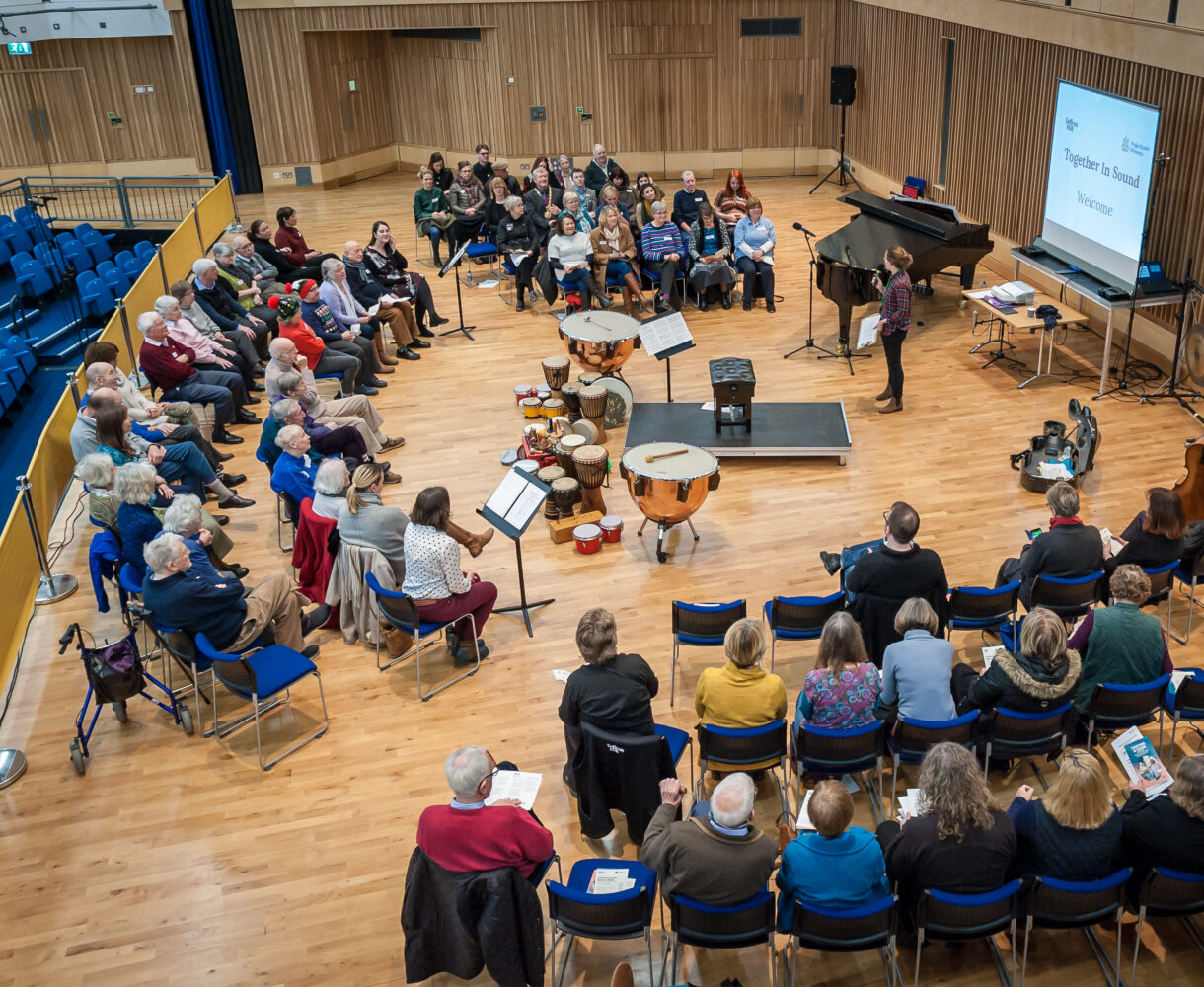 Groups of participants and audience sit in curved seating blocks around a central stage which is full of instruments